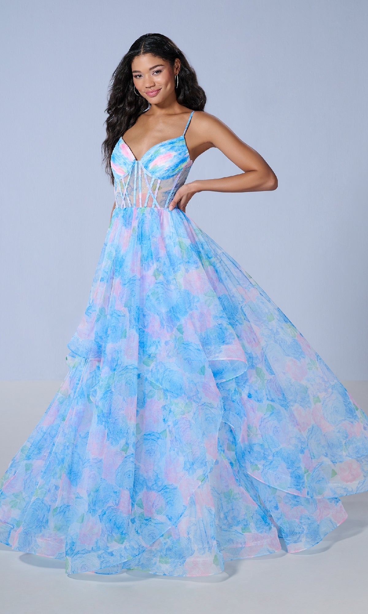 A woman models the Tiffany 16163 long prom dress, featuring pastel blue and pink floral print, thin straps, a sheer corset bodice, and a full layered skirt against a light gray background.